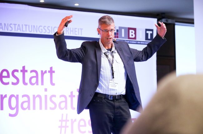 Referent präsentiert auf der IBIT-Fachtagung Veranstaltungssicherheit, umgeben von aufmerksamem Publikum und informativen Grafiken.