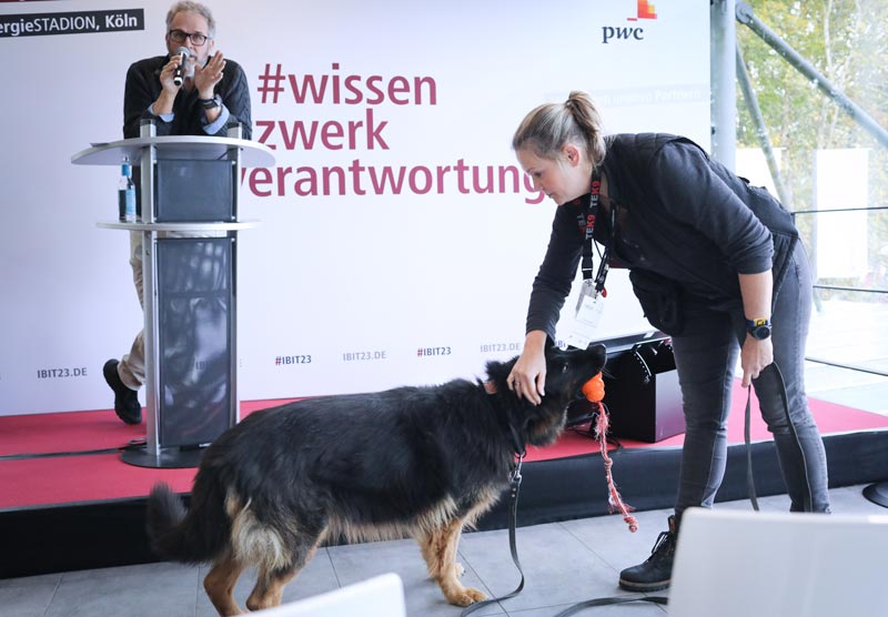 Eine Frau interagiert mit einem Hund auf der Bühne während einer IBIT-Veranstaltung, im Hintergrund ein Redner am Mikrofon.