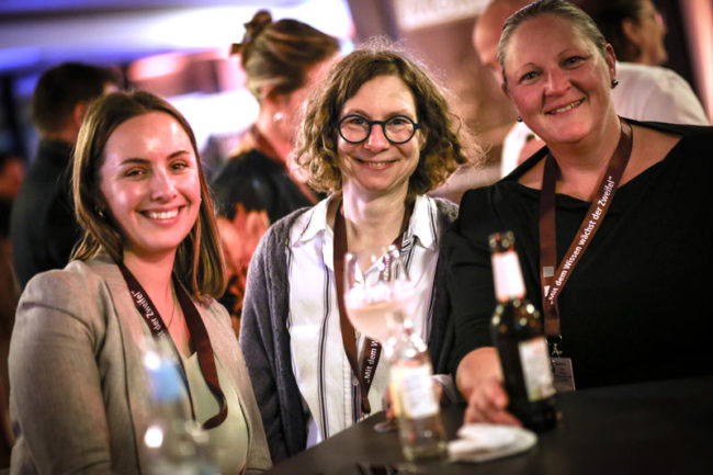 Drei Frauen lächeln entspannt an einem Tisch mit Getränken, umgeben von einer freundlichen, geselligen Atmosphäre beim IBIT.