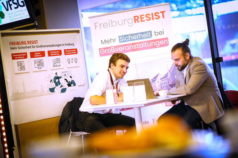 Zwei Personen im Gespräch an einem Tisch, im Hintergrund ein Banner zur Veranstaltungssicherheit. IBIT-Beratung.