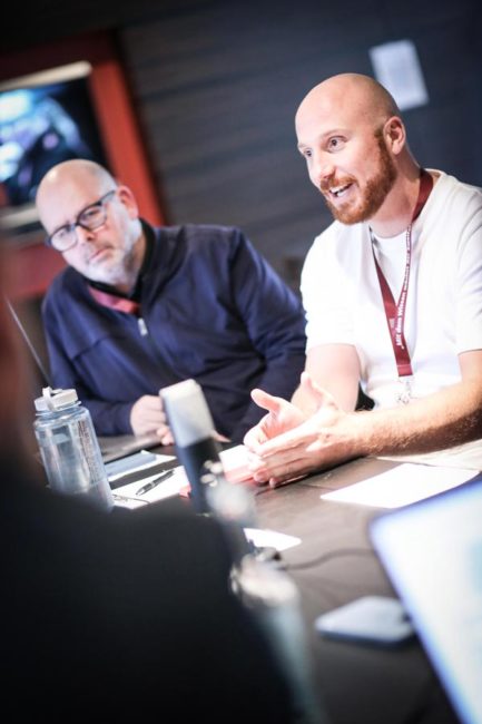 Zwei Männer in einem Besprechungsraum diskutieren lebhaft, während sie an einem Tisch mit Notizen und Wasserflaschen sitzen. IBIT.