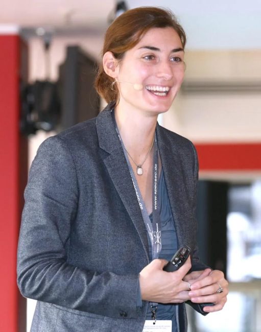 Lächelnde Frau in professioneller Kleidung, die in einem modernen Raum spricht. IBIT-Weiterbildung zum Thema Veranstaltungssicherheit.