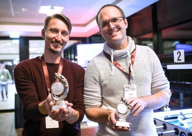 Zwei Männer halten stolz ihre Auszeichnungen in einem modernen Raum, passend zur IBIT-Fachtagung Veranstaltungssicherheit.