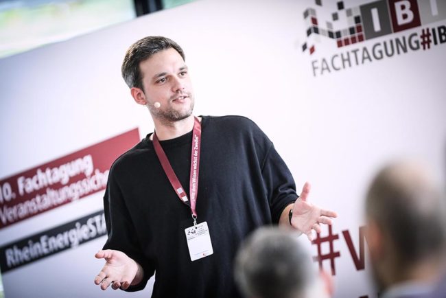 Mann spricht engagiert bei der IBIT-Fachtagung Veranstaltungssicherheit, im Hintergrund Banner und Zuhörer.