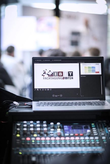 Laptop mit IBIT-Logo und Präsentation im Vordergrund, im Hintergrund unscharfe Teilnehmer bei einer Veranstaltung.