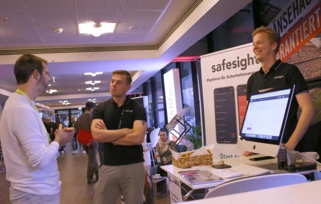 Zwei Personen am Stand eines Unternehmens, das Lösungen für Veranstaltungssicherheit präsentiert. IBIT.