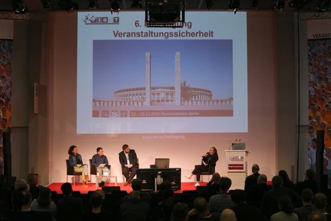 Podiumsdiskussion zur Veranstaltungssicherheit beim IBIT, mit mehreren Sprechern und interessiertem Publikum im Hintergrund.