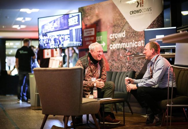 Zwei Männer im Gespräch in einer modernen Umgebung, umgeben von Informationsständen. IBIT-Fachtagung Veranstaltungssicherheit.