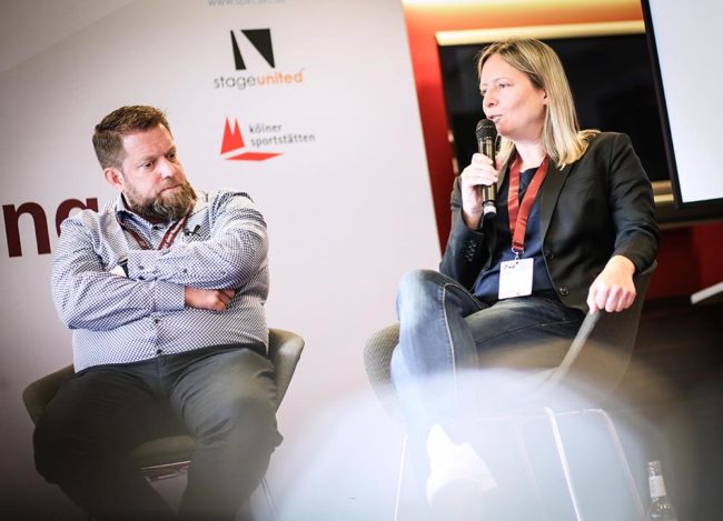 Zwei Referenten diskutieren auf der IBIT-Fachtagung Veranstaltungssicherheit, umgeben von einem modernen Veranstaltungsrahmen.