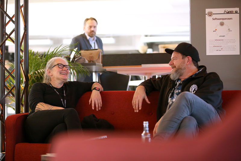 Zwei Personen sitzen entspannt auf einer roten Couch und unterhalten sich, während im Hintergrund ein weiterer Mann beobachtet. IBIT.