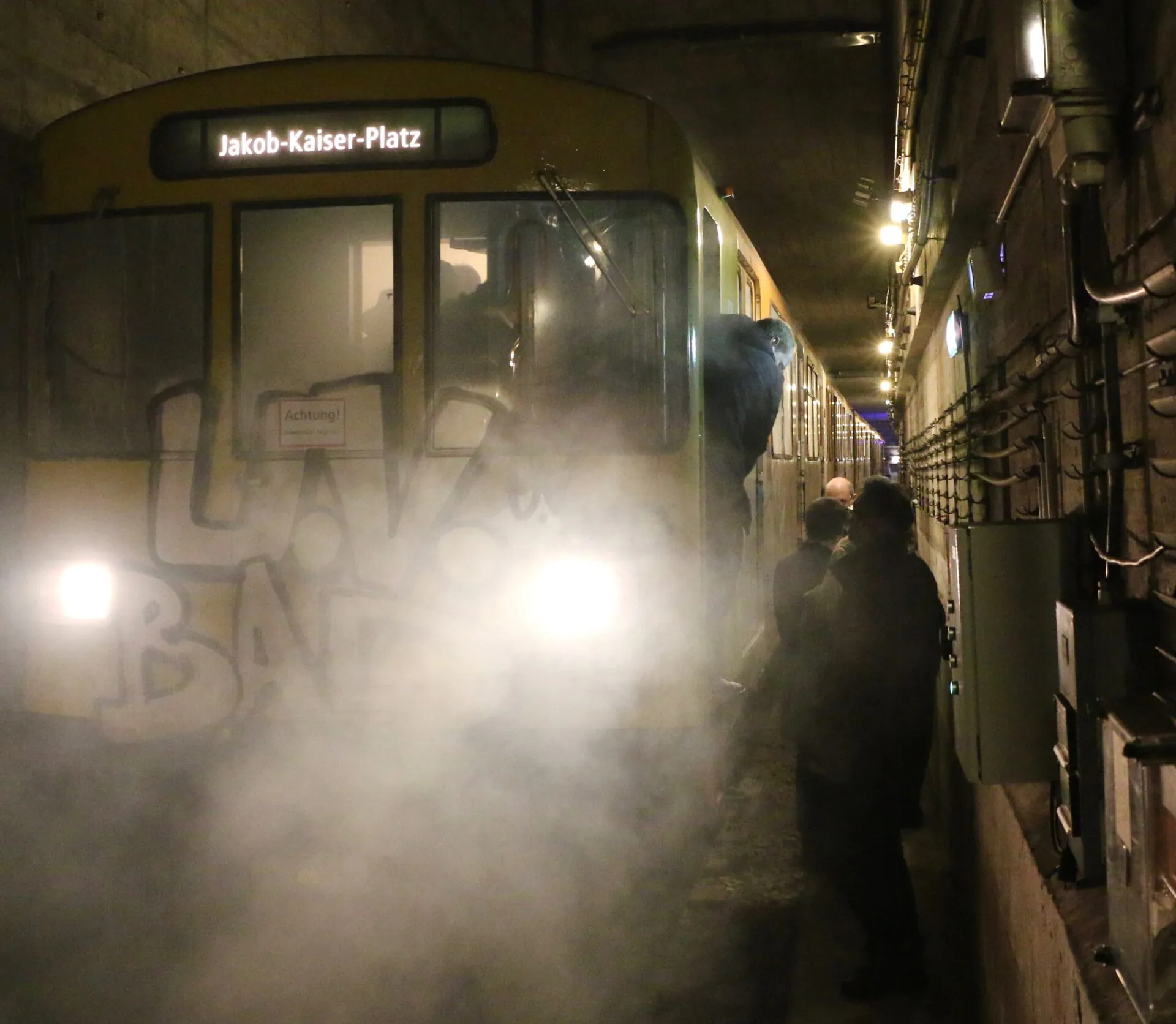 U-Bahn-Station mit dampfendem Zug und Personen im Hintergrund, die auf Sicherheit im öffentlichen Raum achten. IBIT.