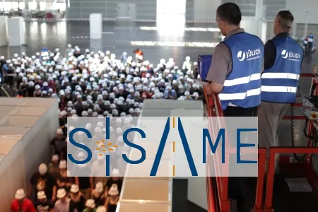 Ein Fachmann beobachtet eine große Gruppe von Teilnehmenden mit Schutzhelmen in einer Veranstaltungshalle. IBIT.