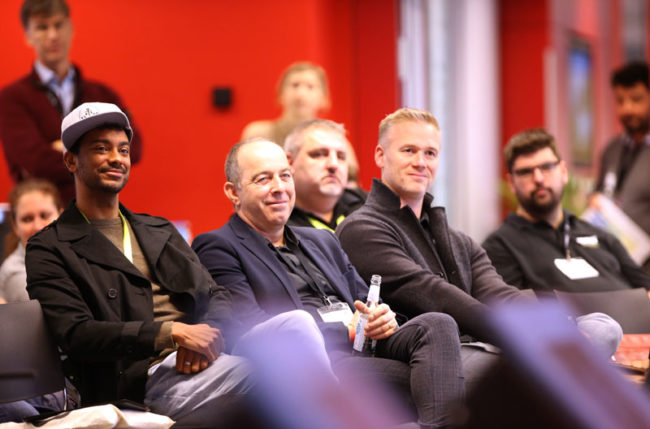 Teilnehmende einer IBIT-Veranstaltung sitzen aufmerksam in einer roten Umgebung und verfolgen die Präsentation auf der Bühne.