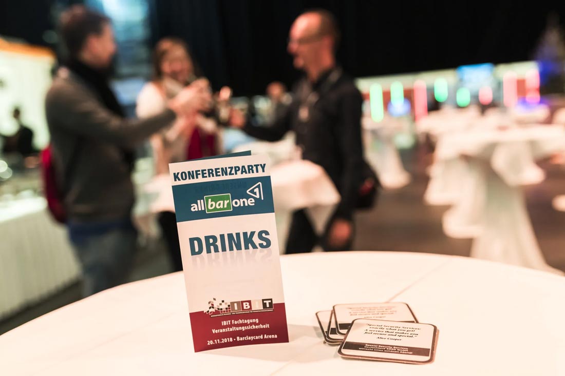 Teilnehmer einer Konferenzparty stehen zusammen und stoßen an, während ein Getränkeständer im Vordergrund sichtbar ist. IBIT Fachtagung Veranstaltungssicherheit.