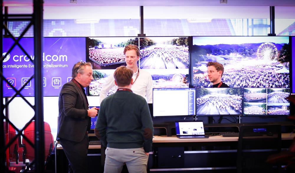 Drei Personen diskutieren an einem Tisch vor mehreren Bildschirmen, die Veranstaltungsszenen zeigen. IBIT.