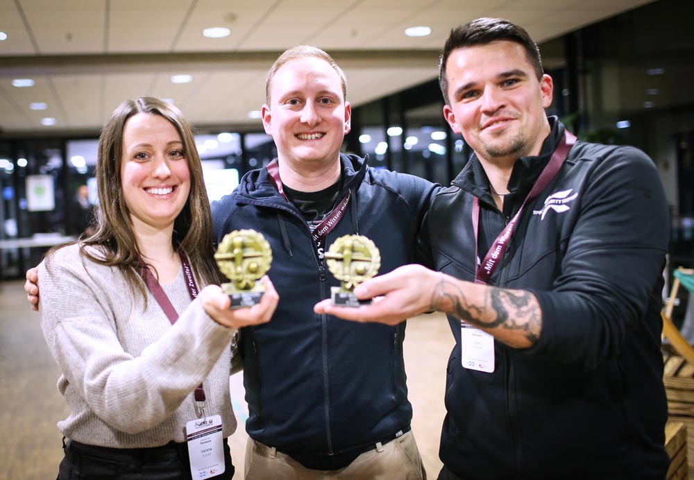Drei Personen halten stolz ihre Auszeichnungen in einer modernen Umgebung, die für eine IBIT-Veranstaltung gestaltet ist.