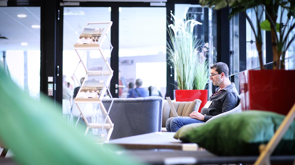 Ein Mann sitzt entspannt in einer modernen Lounge, arbeitet am Laptop. Pflanzen und ein Informationsständer schaffen eine einladende Atmosphäre. IBIT.