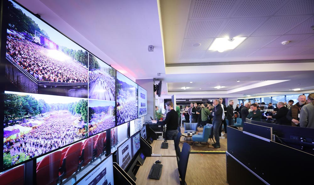 Menschen diskutieren in einem modernen Raum, während große Bildschirme ein Event mit vielen Besuchern zeigen. IBIT.