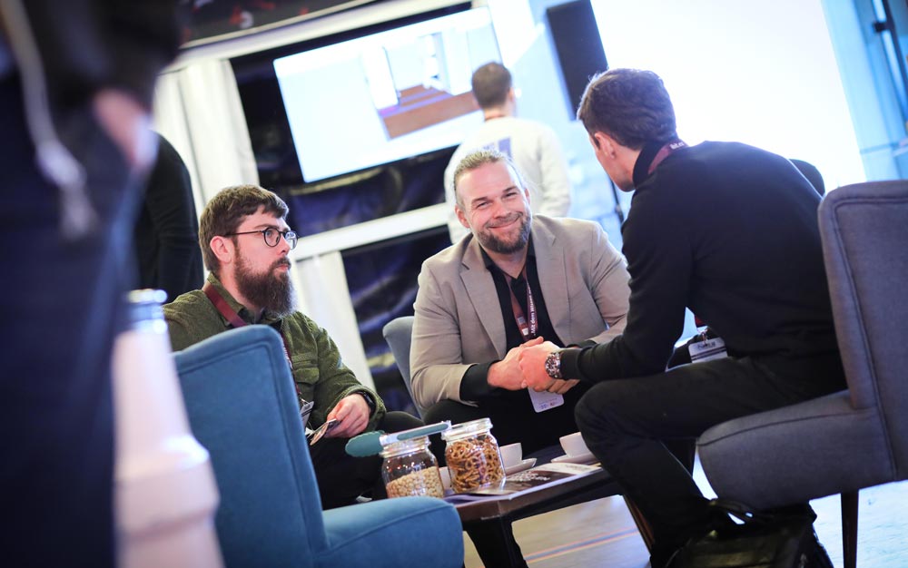 Drei Männer in einem modernen Lounge-Bereich diskutieren und schütteln sich die Hände, während im Hintergrund eine Präsentation läuft. IBIT.